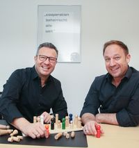 Tobias Kämmerer und Thomas Esche lächeln und vor ihnen steht ein Holzbrett mit vielen Spielfiguren.